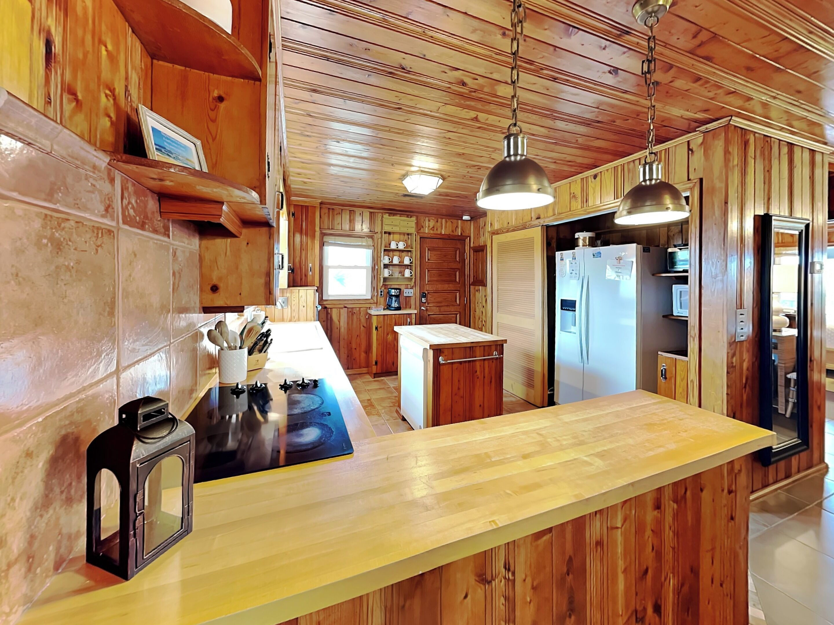 A wooden cabin interior with a kitchen area featuring a central island, a refrigerator, and wooden paneling.