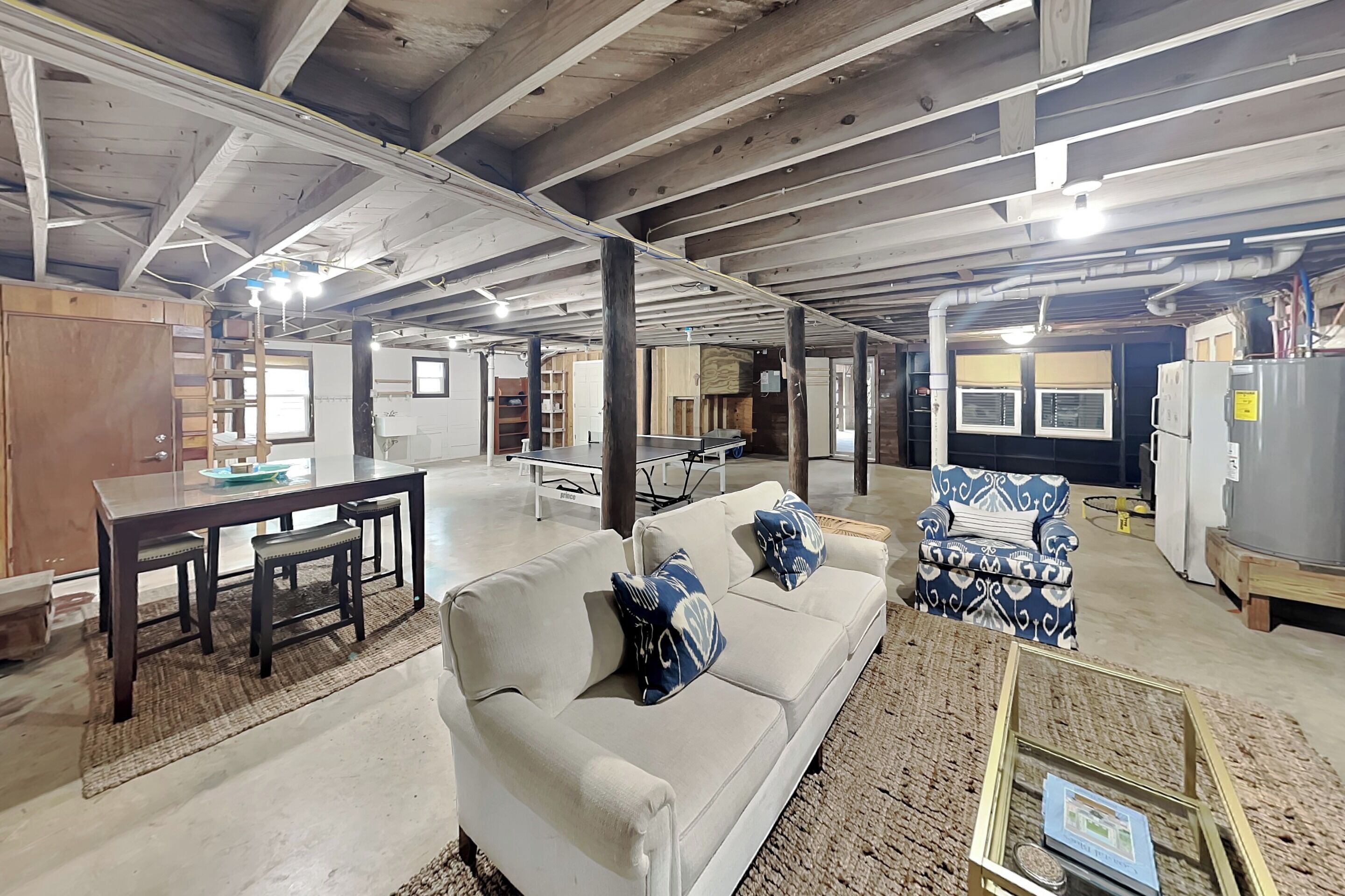 An industrial-style room with exposed beams, a white sofa with blue cushions, a patterned armchair, and a dining table with chairs.