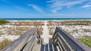 Ein Holzsteg führt zu einem Strand mit Dünen und dem Meer im Hintergrund.