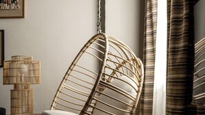 A hanging chair in a room with a wooden floor, a small table with a lamp, and a framed picture on the wall.