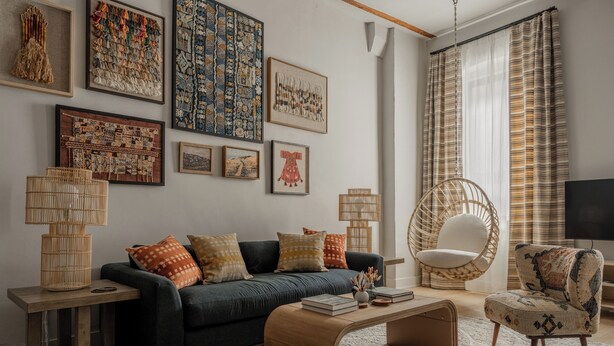 A living room with a dark green sofa, a wooden coffee table, a wicker chair, and a hanging chair. There are framed artworks on the wall and a television on the right.