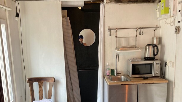 Een kleine keuken met een magnetron, waterkoker en wastafel. Een stoel staat bij de deur, en een prullenbak is zichtbaar.