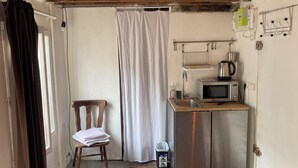 Een kleine keuken met een magnetron, waterkoker en spoelbak. Een stoel staat bij het raam, en een gordijn hangt ernaast.