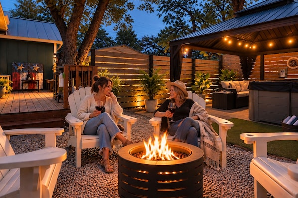 Two people seated by a fire pit in an outdoor patio area with wooden furniture and a brick wall in the background.