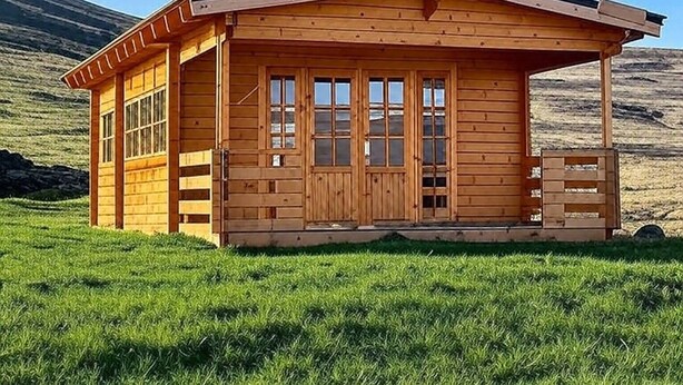 Una cabaña de madera con un porche, ventanas grandes y un campo de césped al fondo.