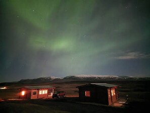 Una cabaña aislada con un coche estacionado afuera, rodeada por un paisaje nevado bajo un cielo nocturno con auroras boreales visibles.