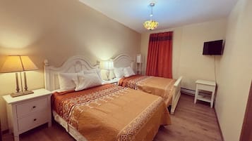 A hotel room with two beds, a nightstand with a lamp, a small table, and a TV mounted on the wall.