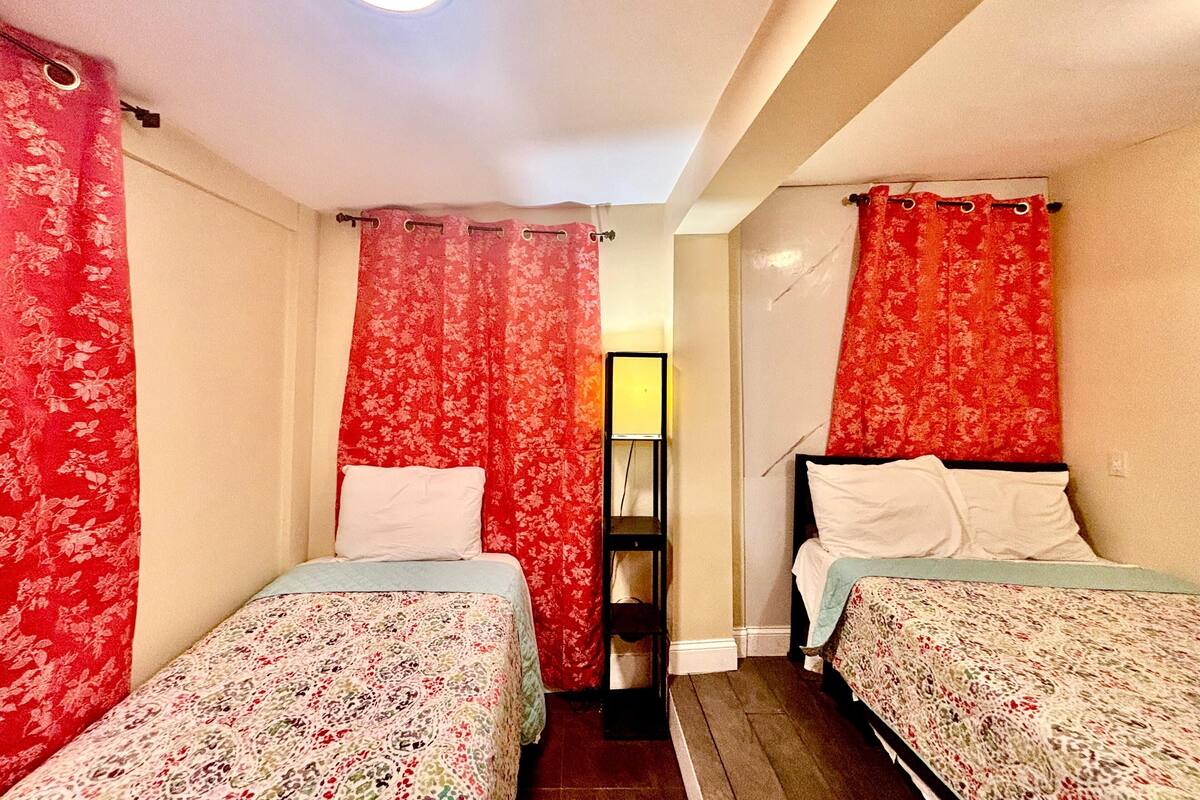 Une petite chambre, de dimensions réduites, avec deux lits simples, chacun agrémenté d’une couvre-lit à motifs floraux et de coussins blancs. La chambre présente un aménagement simple et fonctionnel, avec une petite étagère adossée au mur et un seul luminaire au plafond.