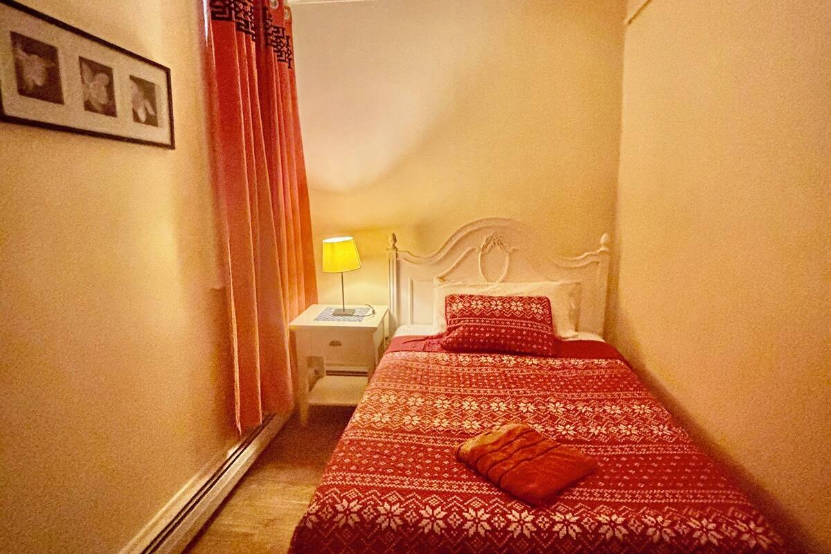 Une petite chambre à coucher, chaleureuse et accueillante, avec un lit simple, une table de chevet, une lampe et un tableau encadré au mur.