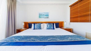 Een netjes opgemaakt bed met een blauw-witte spreideken, twee nachtkastjes met lampen, een houten hoofdbord, een ingelijst foto op de muur en een raam met zonwering.