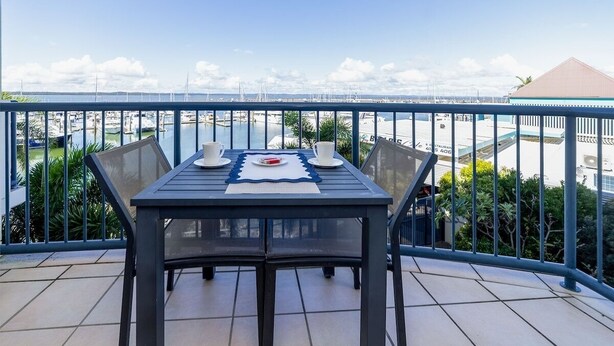 Een balkon met een tafel voor twee, uitkijkend op een jachthaven met boten en een heldere lucht.