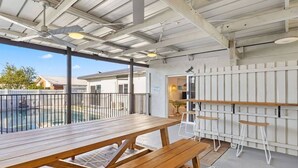 An outdoor patio with wooden tables and chairs, a pool in the background, and a covered area with a ceiling fan.