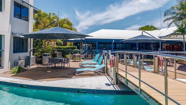 Una piscina con sillones de playa y sombrillas, un área de terraza y un edificio con fachada blanca y detalles azules.