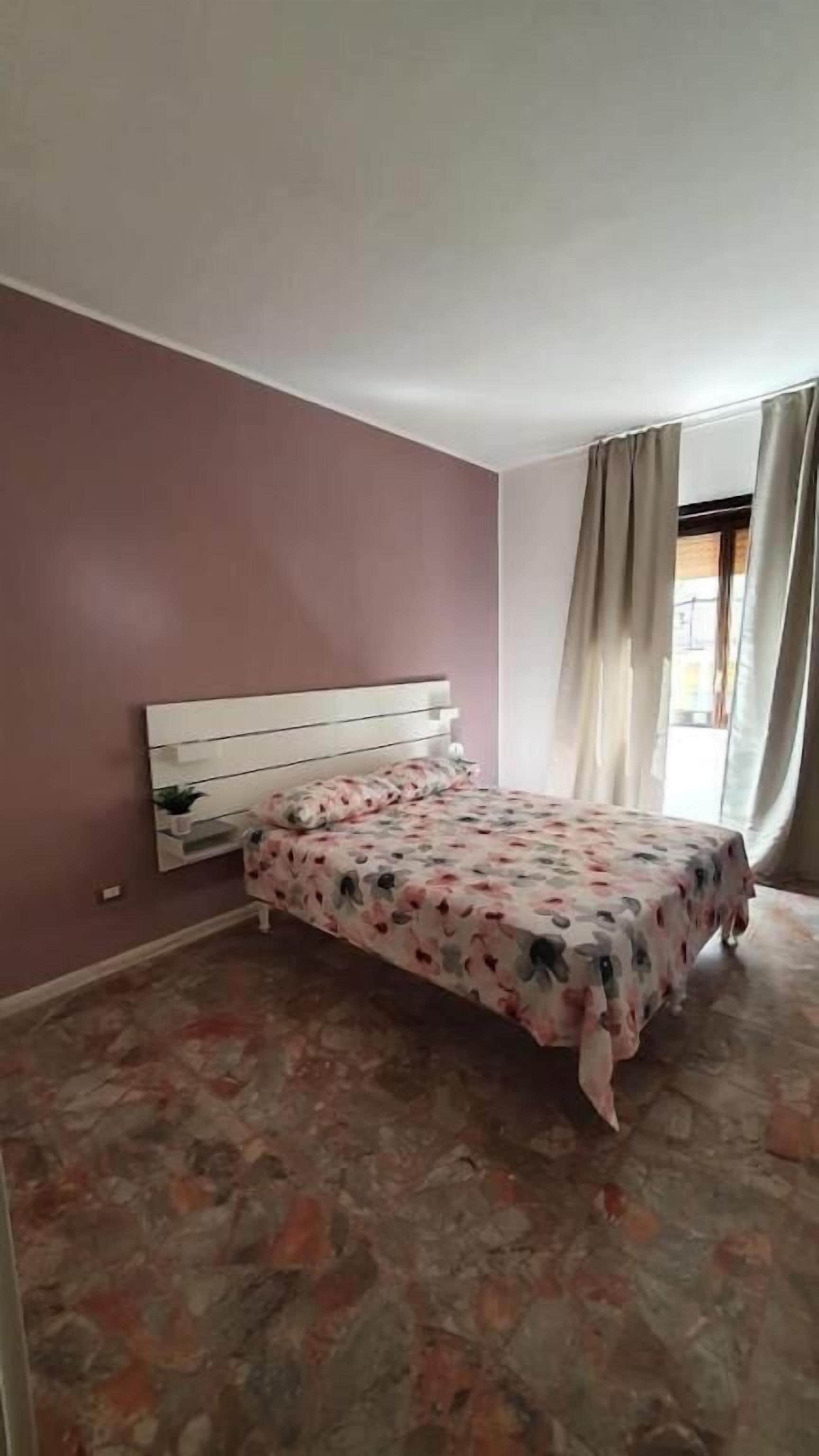 A bedroom with a bed, a nightstand, a window with curtains, and a wall with a textured pattern.