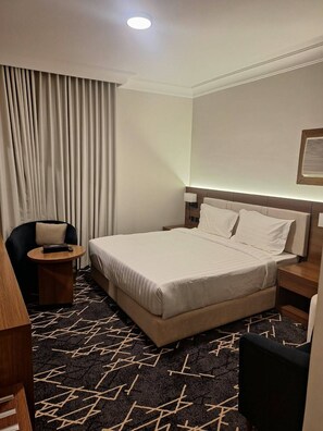 Een hotelkamer met een bed, een stoel, een nachtkastje, een lamp en een raam met gordijnen.