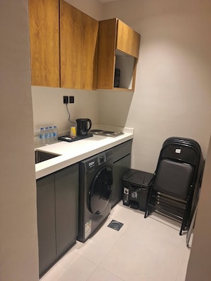 A compact kitchen with a washing machine, a coffee maker, and a stack of black chairs. - Wahat Al Madinah Hotel Apartments (Madinah)