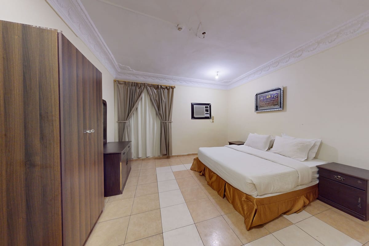 Un dormitorio con cama, armario, mesita de noche, un cuadro enmarco y una ventana con cortinas.