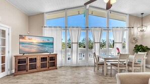A spacious room with a large window, a wooden cabinet with a flat-screen TV, a dining table with chairs, and a ceiling fan. - Bates Avenue House (Indian Rocks Beach)