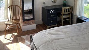 Ein Schlafzimmer mit einem Bett, einem Holzsessel, einem Schminktisch, einem Spiegel, einem Deckenventilator und einer Ziegelwand als Blickfang.