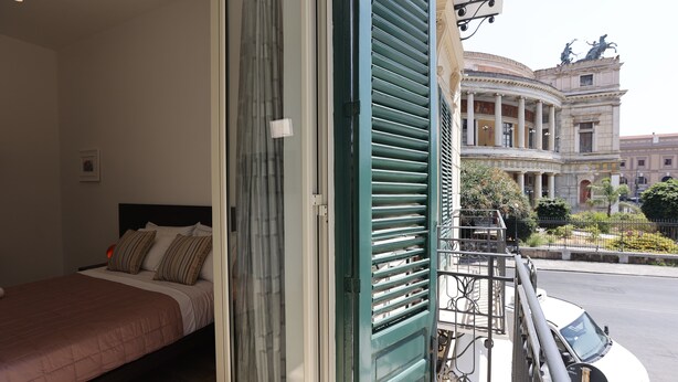 A hotel room with a bed, a window with blinds, and a view of a historic building outside. - Devalù Politeama elegante appartamento al centro (palermo)