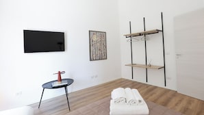 A modern hotel room with a flat-screen TV, a painting, a small table with a book and a red object, a bed with rolled towels, and a wardrobe with a hanging rack. - Devalù Politeama elegante appartamento al centro (palermo)