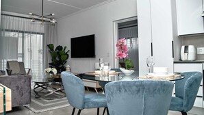 Una sala de estar moderna con comedor, que incluye una mesa de vidrio, sillas tapizadas en azul, una planta grande en maceta y un televisor de pantalla plana montado en la pared.