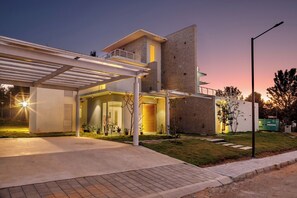 A modern house with a covered driveway, a large brick wall, and a well-lit entrance. - Golf View Home by Stayjade (Hosur)