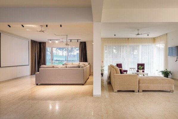 A spacious living area with a large sofa, a smaller sofa, and a coffee table. There are ceiling fans and a projector screen on the wall. - Golf View Home by Stayjade (Hosur)