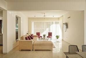 A spacious living room with a beige sofa set, a coffee table, and red armchairs. A ceiling fan is visible, and there is a flat-screen TV mounted on the wall. - Golf View Home by Stayjade (Hosur)