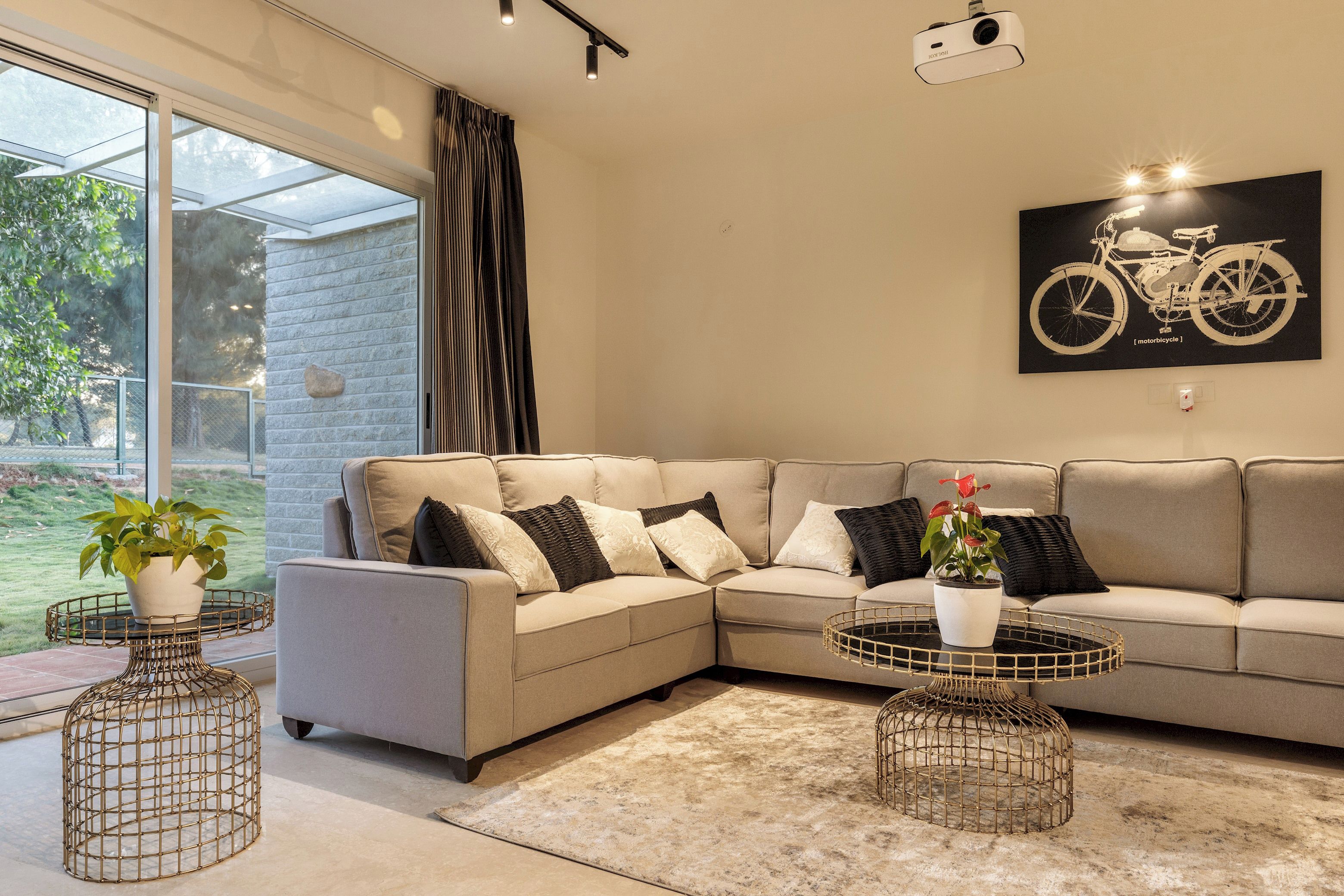 A modern living room with a beige sectional sofa, two round coffee tables with wire bases, and a large window with a view of greenery.