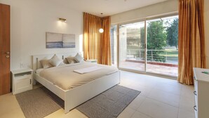 A modern bedroom with a large bed, a ceiling fan, a nightstand, a bedside table, a window with curtains, and a view of the outdoors. - Golf View Home by Stayjade (Hosur)