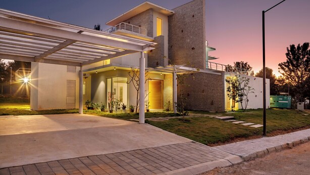 A modern house with a covered driveway, a large brick wall, and a well-lit entrance. - Golf View Home by Stayjade (Hosur)