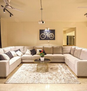 Una sala de estar con un sofá seccional color beige, una mesa redonda para café, un cuadro enmarcado de una motocicleta y un proyector instalado en el techo.