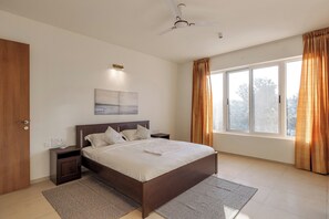 Un dormitorio con una cama grande, una mesita de noche, un ventilador de techo, una ventana con cortinas y un cuadro en la pared.