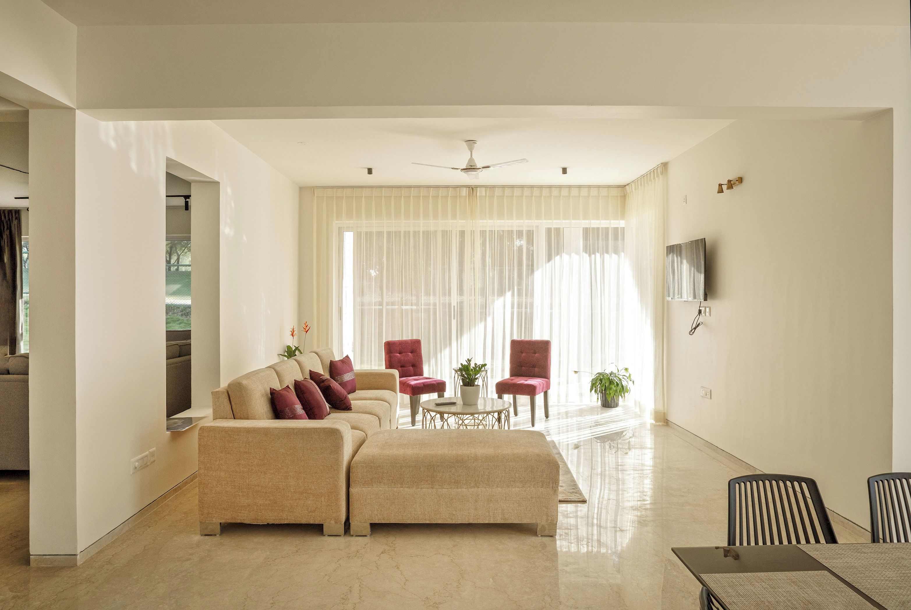 A spacious living room with a beige sofa set, a coffee table, and red armchairs. A ceiling fan is visible, and there is a flat-screen TV mounted on the wall.