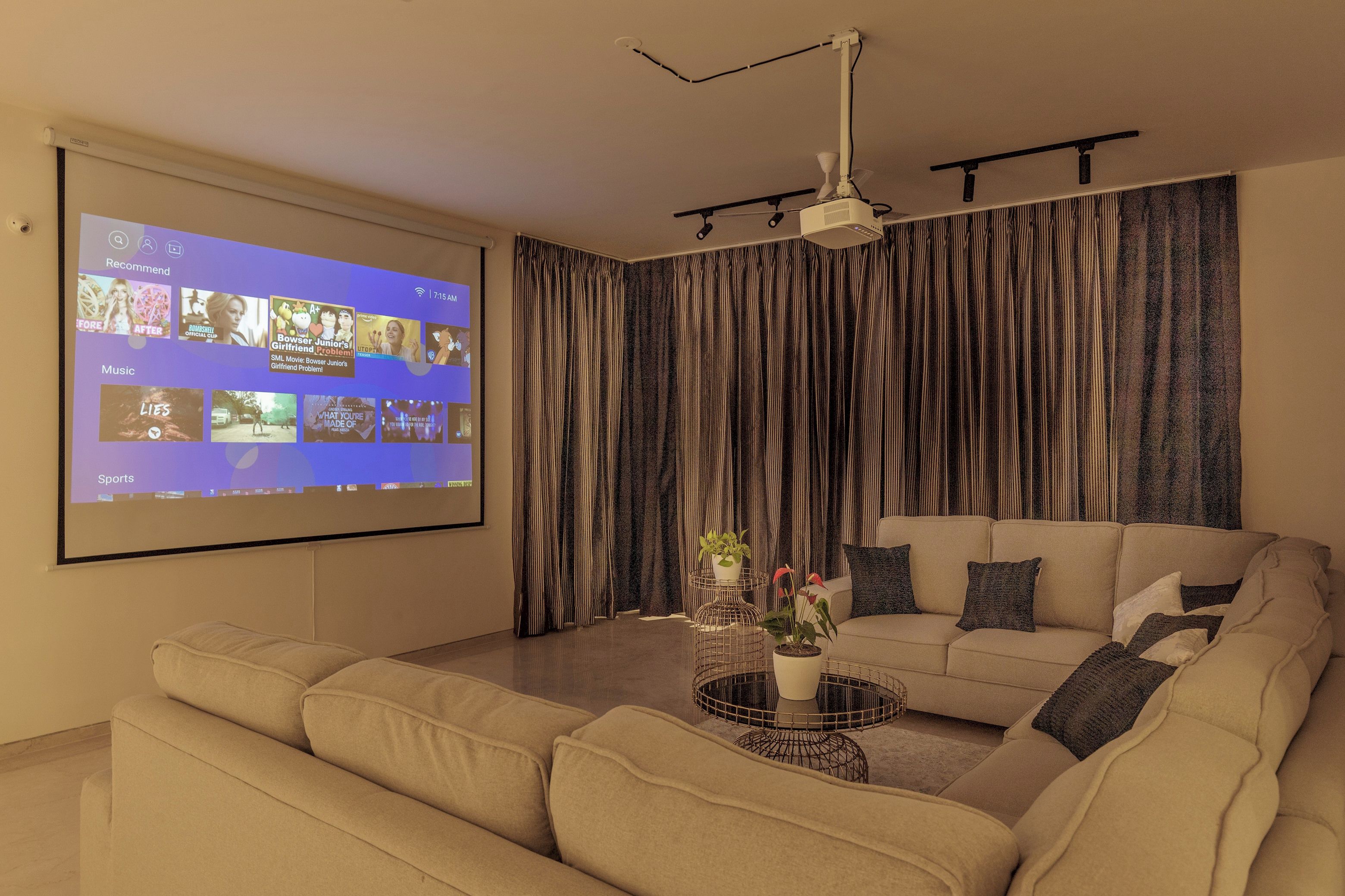 A modern living room with a large screen displaying a streaming service interface, a beige sofa, a coffee table with plants, and a ceiling-mounted projector.