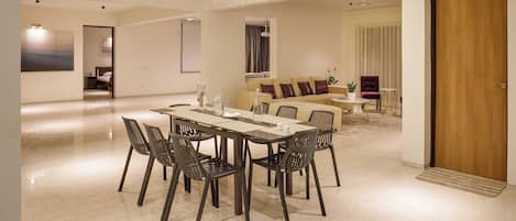 A spacious living area with a dining table, chairs, and a sofa. There are ceiling fans, a large window with curtains, and a door.