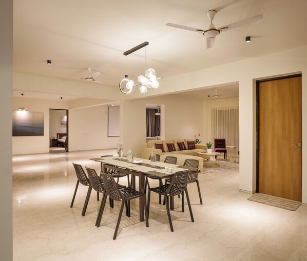 A spacious living area with a dining table, chairs, and a sofa. There are ceiling fans, a large window with curtains, and a door. - Golf View Home by Stayjade (Hosur)