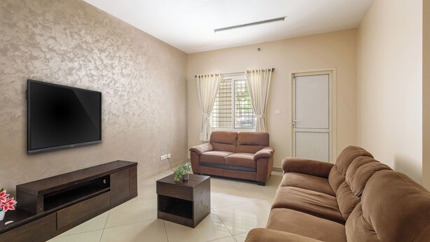 A living room with a flat-screen TV mounted on the wall, a brown sofa, a coffee table with a plant, and a door with a window. - Aspire Heights by Red Olive (Bengaluru)