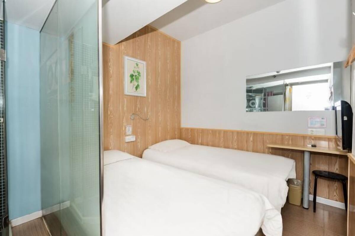 A hotel room with two beds, a wooden wall panel, a small desk, a mirror, and a glass shower enclosure.