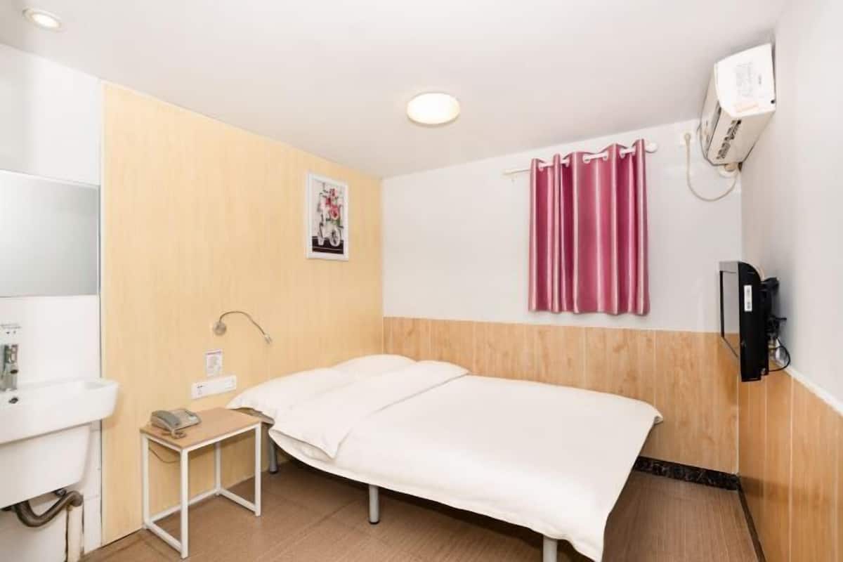 A hotel room with a bed, a small table with a phone, a wall-mounted TV, and a window with curtains.