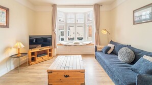 Sala de estar com sofá, televisão, mesa de centro de madeira e janela com cortinas.
