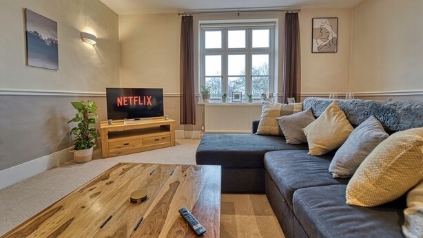 A living room with a sectional sofa, a wooden coffee table, a TV on a stand, and a potted plant. - Harrow Croft, 2 Bedroom Flat St Johns, Worcester (Worcestershire)