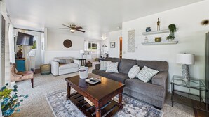 Uma sala de estar ampla, com sofá, mesa de centro e televisão. Há prateleiras com objetos decorativos e um ventilador de teto.