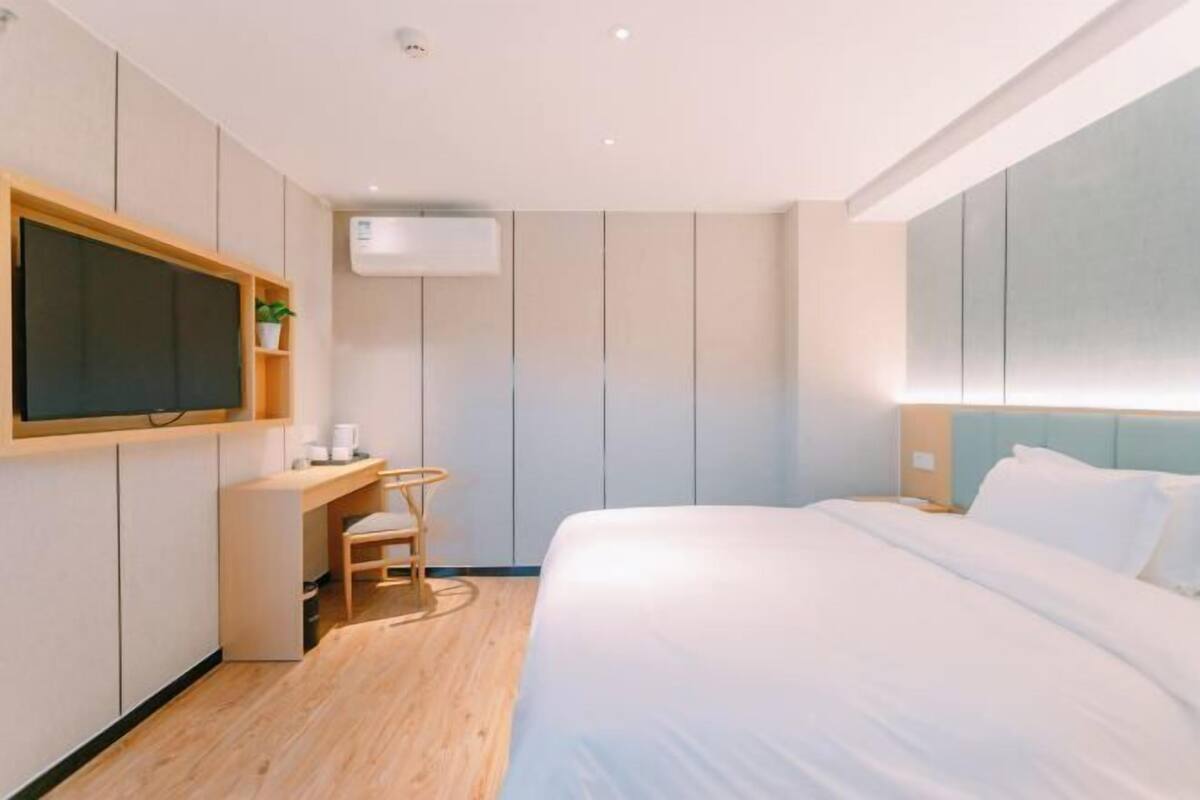 A modern hotel room with a bed, a desk with a chair, a television, and an air conditioning unit.