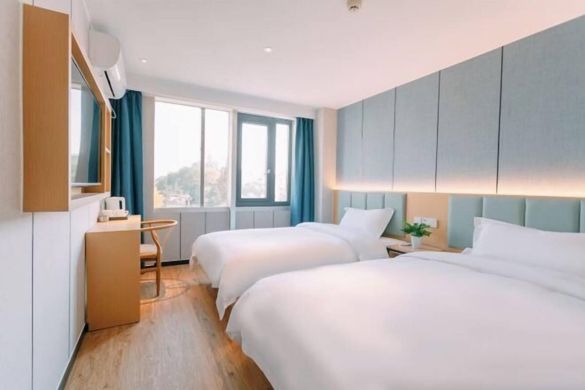 A hotel room with two beds, a desk, a chair, a window with curtains, and a view of the city.