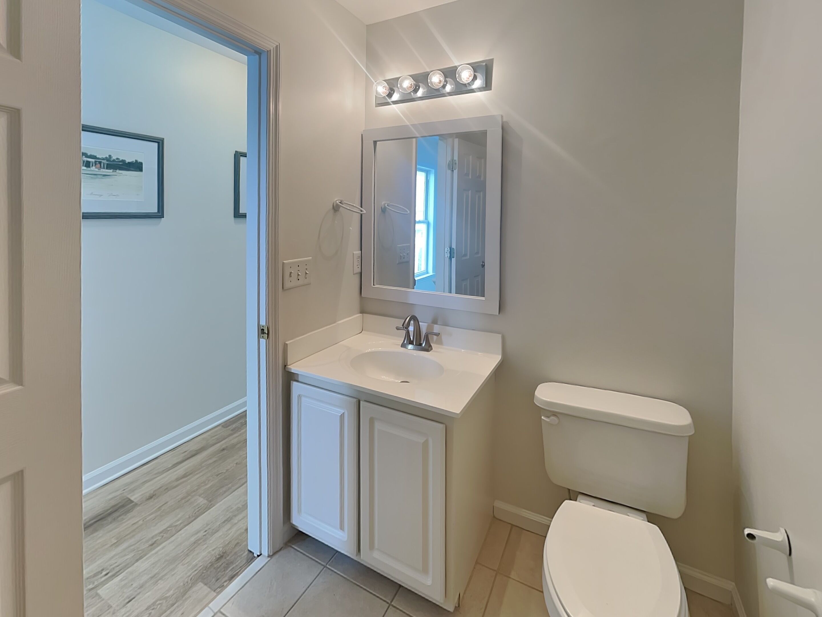 Een badkamer met een witte wastafel, meubel en toilet. Er hangt een lijst met een afbeelding aan de muur en een deur die naar een andere kamer leidt.