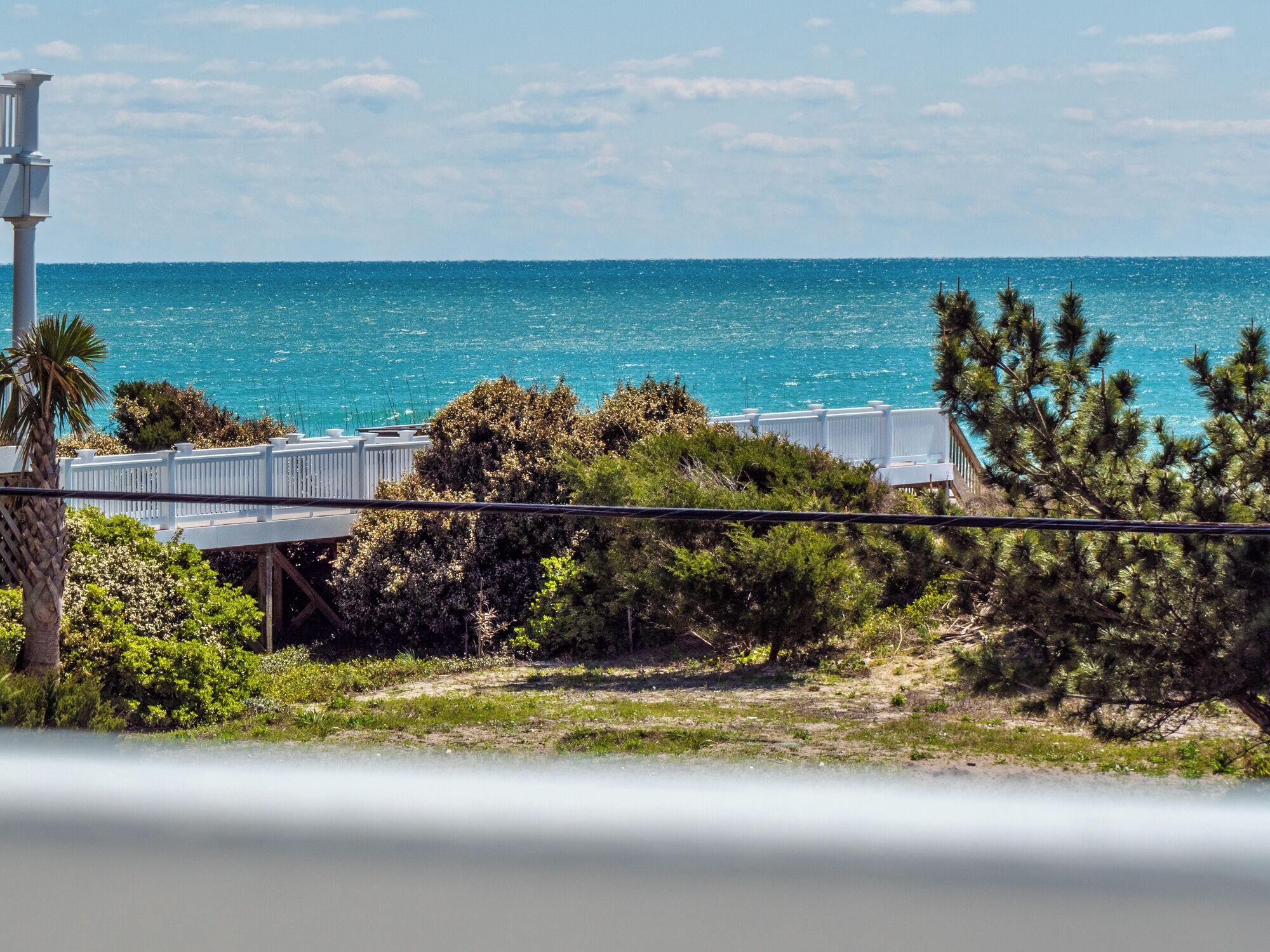 A coastal view with a white fence, greenery, and a clear blue sea.