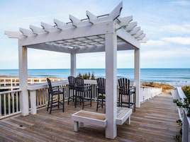 Um deck de madeira com pérgola, cadeiras e um banco com vista para o oceano.