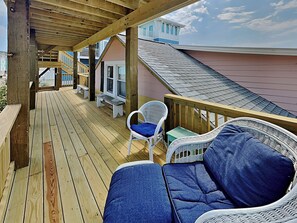 A wooden deck with a covered seating area, including a wicker chair and a sofa with blue cushions. - Beach Breeze by Carolina Retreats (Carolina Beach)